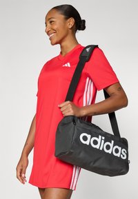 Černá adidas taška přes rameno z tkaniny, s výrazným bílým logem, zipovým uzávěrem a nastavitelným ramenním popruhem, kterou drží osoba.