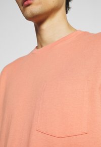 Plan rapproché d'une personne portant un t-shirt à col rond de couleur saumon avec une poche poitrine cousue sur le côté gauche.