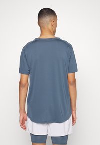 Tmavě modré sportovní tričko s krátkým rukávem a kulatým výstřihem, hladký materiál, mírně prodloužený zadní lem, nošeno s světlými šortkami.