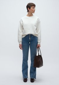 Krém színű pulóver, magas derekú kék szárszélesítő farmer, valamint egy texturált barna kézitáska. A modell rövid hajú, sötét cipőket visel.