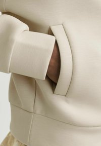 Sudadera beige con un bolsillo lateral de apertura curva, hecha de una tela suave con una textura delicada y un diseño minimalista.