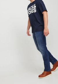 Navy sininen t-paita, jossa on valkoiset tekstit "JACK & JONES," yhdistettynä sinisiin farkkuihin ja ruskeisiin nauhakenkiin, joissa on valkoiset pohjat.