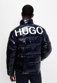 Mørkeblå pufferjakke med en blank finish, prydet med store hvide "HUGO"-bogstaver på ryggen, horisontalt quiltet mønster og høj krave.