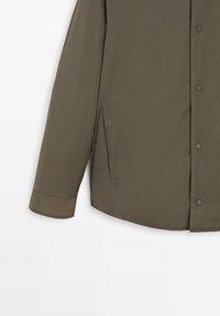 Chemise boutonnée vert olive, avec un col droit, des manches longues, une texture de tissu lisse, des fentes latérales et des pressions.