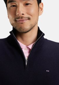 Pull en laine bleu marine zippé avec un col haut, superposé sur une chemise polo rose pâle. Doté d'une fermeture éclair argentée et d'un petit logo sur la poitrine gauche.