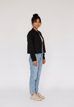 Giovane donna con capelli scuri raccolti in uno chignon, indossa una giacca nera, una maglietta bianca, jeans azzurri e sneakers beige, in piedi su sfondo bianco.