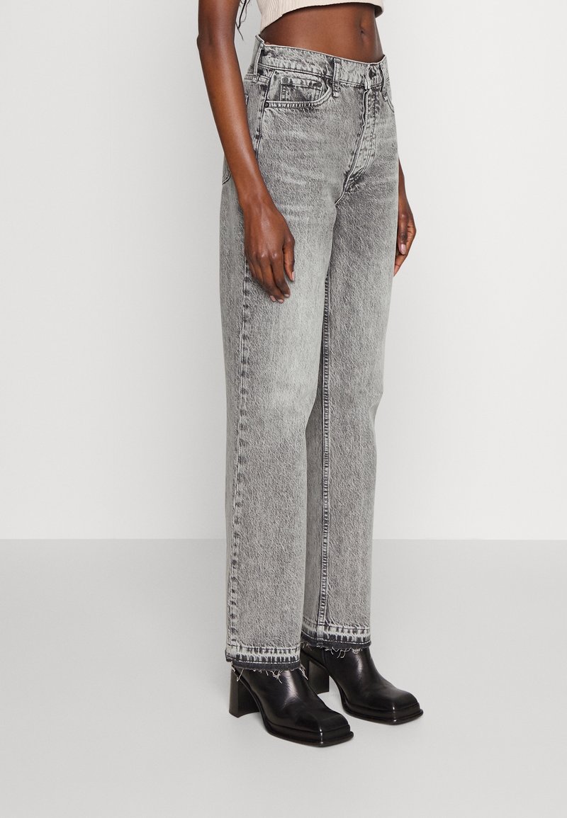 Femme portant un jean gris délavé à l'acide, taille haute et jambes droites, avec un ourlet brut, associé à des bottines à talons en cuir noir.