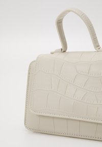 Torebka w beżowej skórze z wzorem krokodyla, o strukturalnym kształcie i górnym uchwycie. Szczegółowe przeszycia dodają faktury powierzchni.