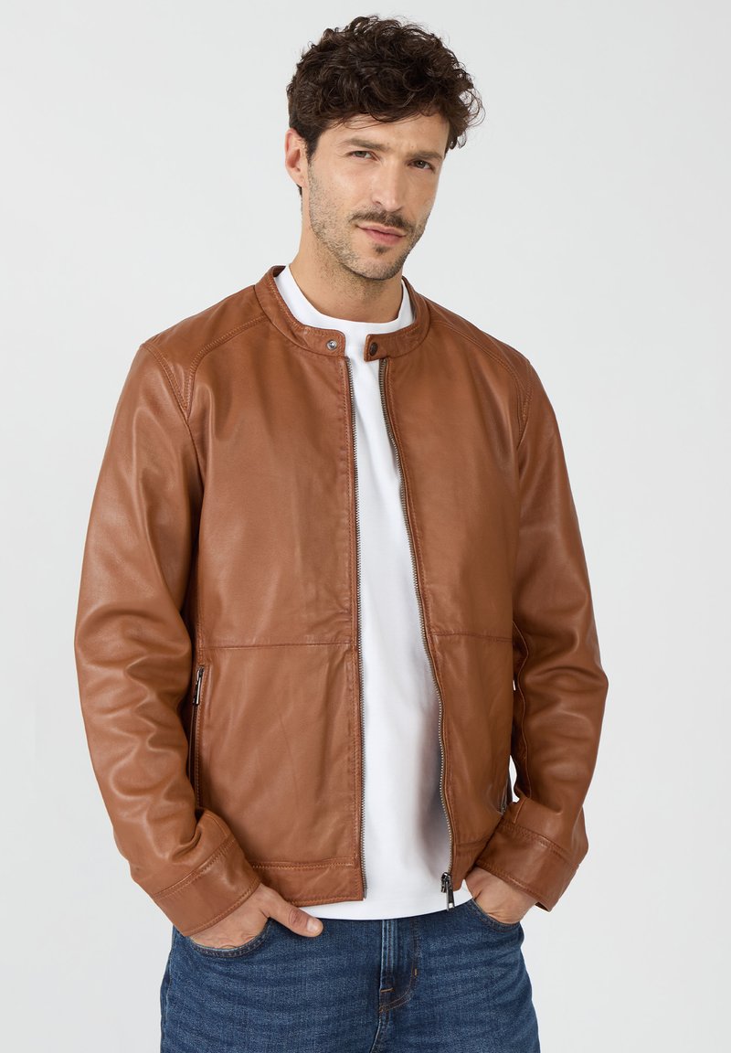 Braune Lederjacke mit rundem Kragen, Reißverschluss vorne und seitlichen Reißverschlusstaschen. Glatte Textur und taillierte Passform. Kombiniert mit einem weißen Hemd.