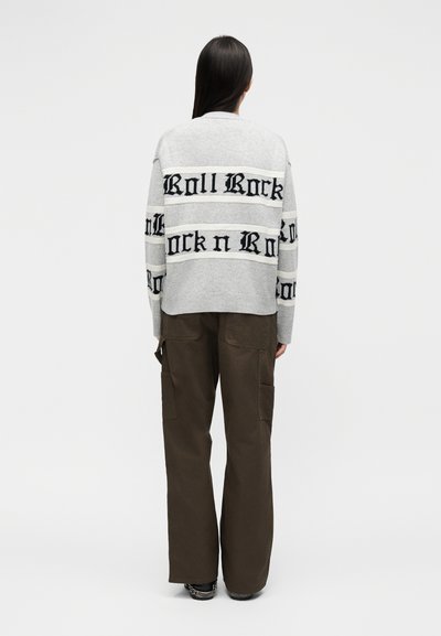 Άτομο με μακριά σκούρα μαλλιά που φοράει ανοιχτό γκρι πουλόβερ με το κείμενο «Rock n Roll» και σκούρο καφέ παντελόνι φαρδιάς γραμμής, όρθιο, γυρισμένο προς τα πίσω.
