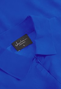 Modrá blúzka s hladkou textúrou, s golierom, predným zipsom a čiernou nálepkou s nápisom "Vascha STOCKHOLM" v bielej farbe. Veľkosť S.