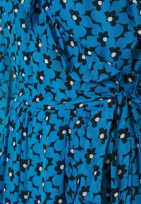 Vestido azul con flores que presentan flores negras con centros blancos, detalles con volantes y una cintura atada. Hecho de una tela ligera con pliegues.