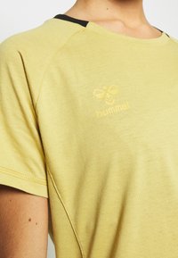 Kollane lühikeste varrukatega särk nähtavate õmblustega, mida kannab inimene, millel on väike mesilase logo ja tekst "hummel" rinnal.