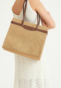 Tan woven tote bag with a textured finish, featuring brown leather handles and trim. The design is rectangular, with a structured shape.