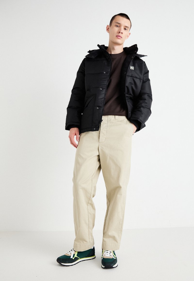 Chaqueta de plumas negra con cierre de cremallera y botón a presión, pantalones beiges y zapatillas verdes con acentos amarillos. El modelo está de pie frente a un fondo plain.