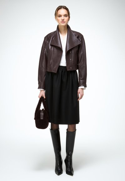 Nő barna bőrkabátot, fehér blúzt, fekete midi szoknyát és fekete, térdig érő csizmát visel, kezében barna textúrált kézitáskával, áll.