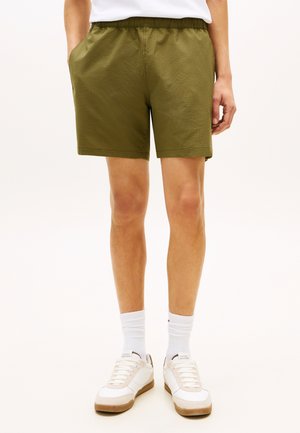 Olívazöld texturált rövidnadrágot, fehér térdzoknit és fehér tornacipőt viselő személy, aki részben zsebre tett kézzel áll.