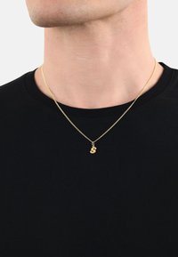 Collana d'oro con un pendente a forma di "s" minuscola su una catena sottile e delicata, indossata sopra una maglietta nera. La catena presenta una fine texture.