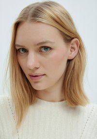 Nahaufnahme einer Person mit glattem, schulterlangem blonden Haar und blauen Augen, die einen cremefarbenen Strickpullover mit geripptem Muster trägt.