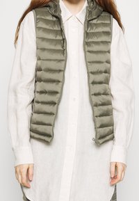 Zateplená vestuška olivové barvy s kapucí, zipovým uzávěrem a vertikálním prošíváním, nošená přes světle zbarvenou košili na knoflíky.