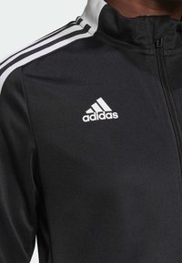 Černá sportovní bunda s vysokým límcem, zapínáním na zip, třemi bílými pruhy na ramenou a vyšitým bílým logem Adidas na hrudi.