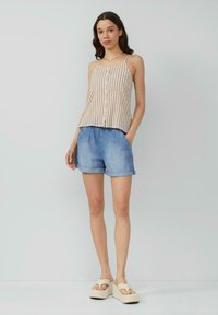 Lichtgewicht beige gestreepte tanktop met knopen, gecombineerd met denim shorts. Model draagt beige platform sandalen en staat tegen een neutrale achtergrond.