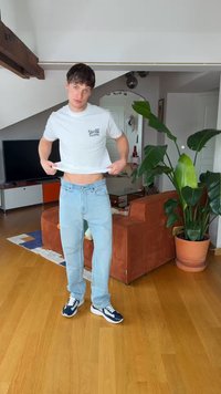 Ung mand løfter hvid T-shirt for at vise maven, iført lyseblå jeans og sneakers, stående i stuen med en plante og orange sofa.