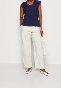 Námořnický volánkový top s výstřihem do V, stažený v pase; spárováno s krémovými širokými nohavicemi a bílými slingback botami se zlatými akcenty.