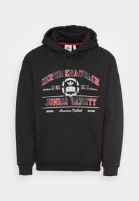 Sweat-shirt à capuche noir avec le texte rouge et blanc "Herzogenaurach Junior Varsity", le logo Adidas et une poche kangourou à l'avant.
