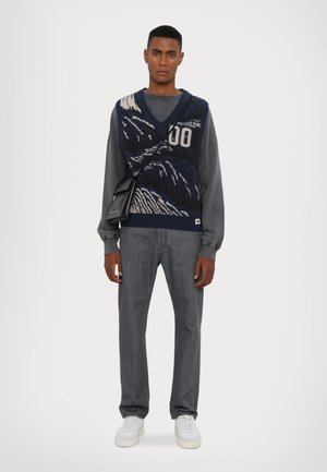 Marineblauer, gestrickter Sweater-Body mit Wellenmuster, weißen Akzenten, über einem grauen Sweatshirt getragen, kombiniert mit grauen Jeans und weißen Sneakers.