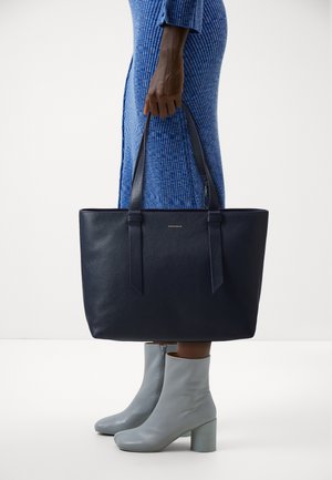 Navy leather tote bag with textured finish, vertical seams, and long handles. Paired with light gray ankle boots and a blue striped dress.