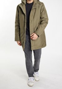 Oliven grøn vandtæt parka jakke med hætte, trykknaplukninger og justerbare snore, parret med grå bukser og hvide sneakers.