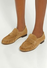 Tan suede slip-on loafers with a rounded toe, raised seam detailing, and a wide band across the upper for added design interest.