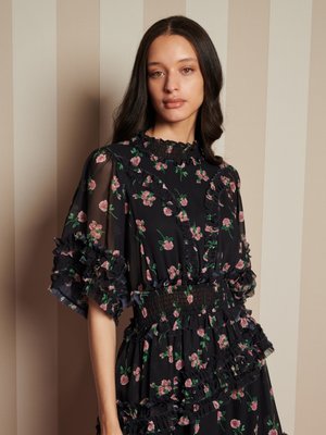 Jovem mulher com cabelo longo e escuro veste um vestido floral preto com mangas transparentes ruffled e cintura franzida, em frente a uma parede listrada em bege.