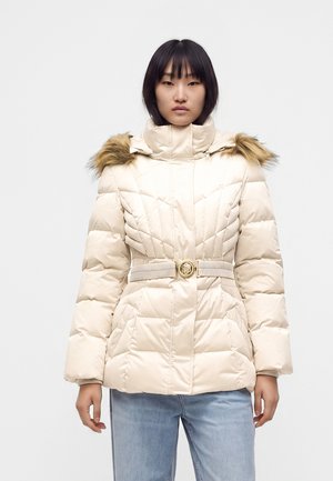 Femme portant une veste matelassée crème avec bordure en fourrure sur la capuche, taille cintrée avec ceinture, et jean bleu clair, debout devant un fond clair uni.