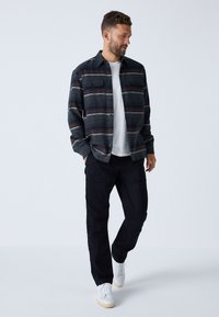 Camicia a quadri nera e grigia con bottoni, maniche lunghe, indossata sopra una t-shirt bianca, abbinata a pantaloni cargo neri e sneaker bianche.