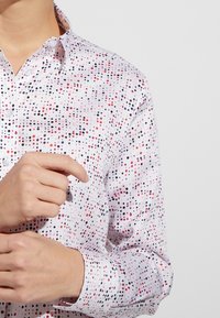 Persona che indossa una camicia bianca a maniche lunghe con un motivo a pois multicolori mentre aggiusta il polsino del braccio sinistro.