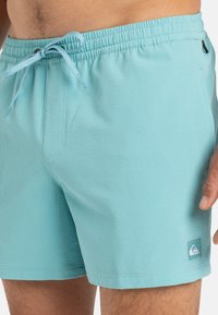 Svetle modré plavky s texturovanou látkou, elastickým pasem, nastavitelnou šňůrkou a malou nášivkou loga na dolní části nohy.