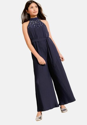 Dievča v tmavomodrom plisovanom overal bez rukávov s ozdobným výstrihom a striebornými balerínami, stojace s uvoľnenými rukami po bokoch.