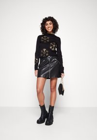 Schwarzer Pullover mit goldenen Schneeflockenmuster, kombiniert mit einem schwarzen Leder-Minirock mit vorderen Reißverschlüssen und Stiefeletten. Hält eine schwarze Tasche.