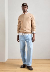 Felpa color cammello con scollatura rotonda, indossata sopra una maglietta a righe. Jeans azzurri chiari, scarpe marroni e un cappello marrone. Dettagli del logo minimi.