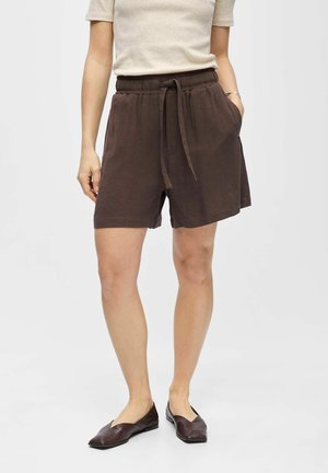 Žena v hnedých šortkách so šnúrkou, béžovom tričku s krátkym rukávom a tmavohnedých plochých topánkach, stojaca s jednou rukou v vrecku.