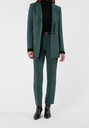 Femme portant un costume à motifs turquoise avec blazer cintré, pantalon assorti, col roulé noir, ceinture et bottines noires.