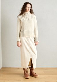 Suéter de punto beige con cuello alto, dobladillo y puños acanalados, combinado con una falda midi color crema que cuenta con una abertura frontal y botas marrones con cordones.