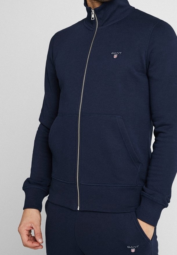 Homme portant une veste zippée bleu marine et un pantalon assorti avec le logo GANT sur la poitrine et la cuisse, mains détendues sur les côtés.