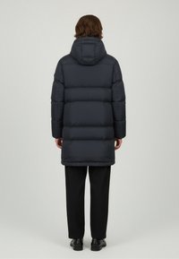 Persona de espaldas vestida con un abrigo largo y acolchado negro con capucha, pantalones negros y zapatos negros, de pie frente a un fondo liso.