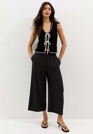 Kobieta o długich ciemnych włosach, ubrana w czarny bezrękawnik z białymi wiązaniami oraz czarne spodnie o szerokich nogawkach, stojąca z rękami w kieszeniach.