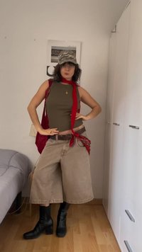 Junge Person in braunem Tanktop, lockeren beigefarbenen Shorts, schwarzen Stiefeln, rotem Schal und Bandana sowie Tarnkappe posiert drinnen mit Händen in der Hüfte.