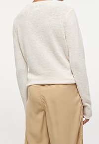Pull à manches longues blanc texturé avec un col rond, associé à un pantalon beige. Le tissu présente un motif tissé et un léger éclat.