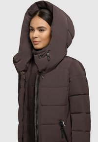 Giacca imbottita marrone con un grande cappuccio sagomato, chiusure con zip e bottoni a pressione e tasche laterali con zip. Presenta una texture trapuntata e un design elegante.